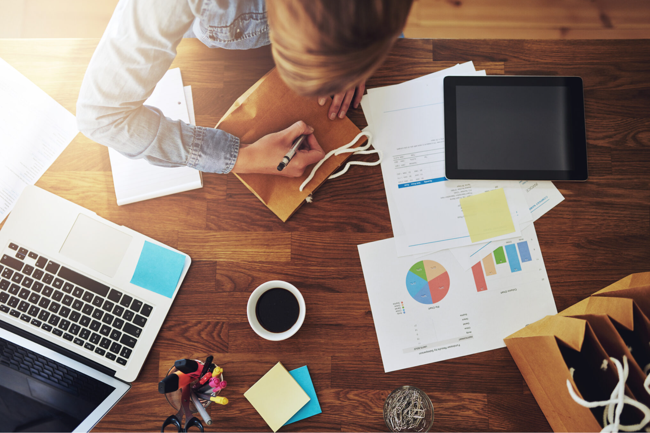 A woman on a desk working with papers, spreadsheets and a coffee around