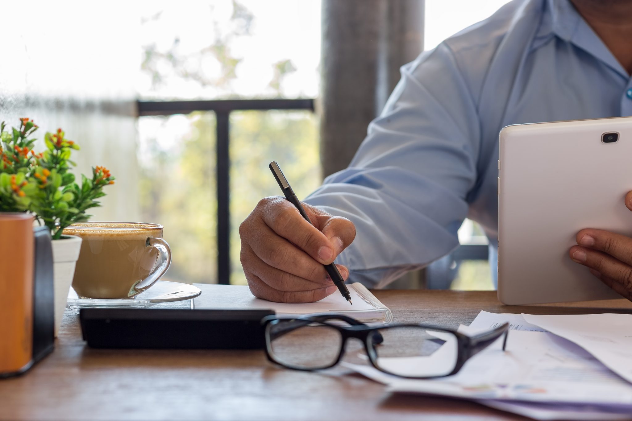 Un homme écrivant sur un cahier avec une tasse de café et ses lunettes à ses côtés.